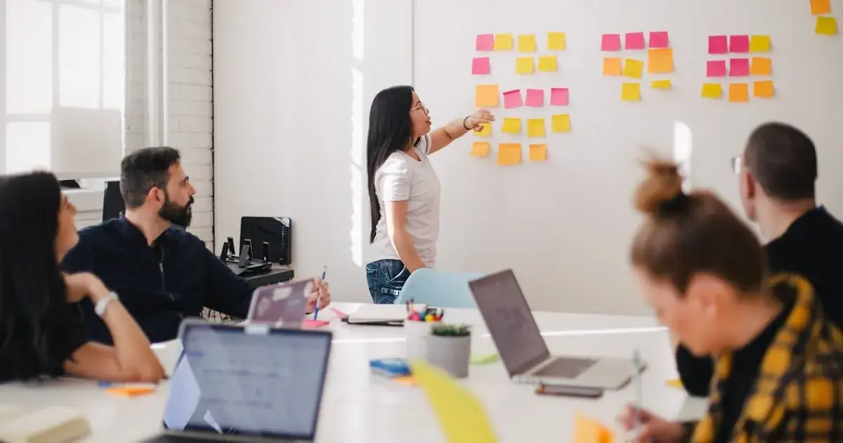 A woman points at pink and yellow sticky notes on a whiteboard while colleagues work at laptops nearby.