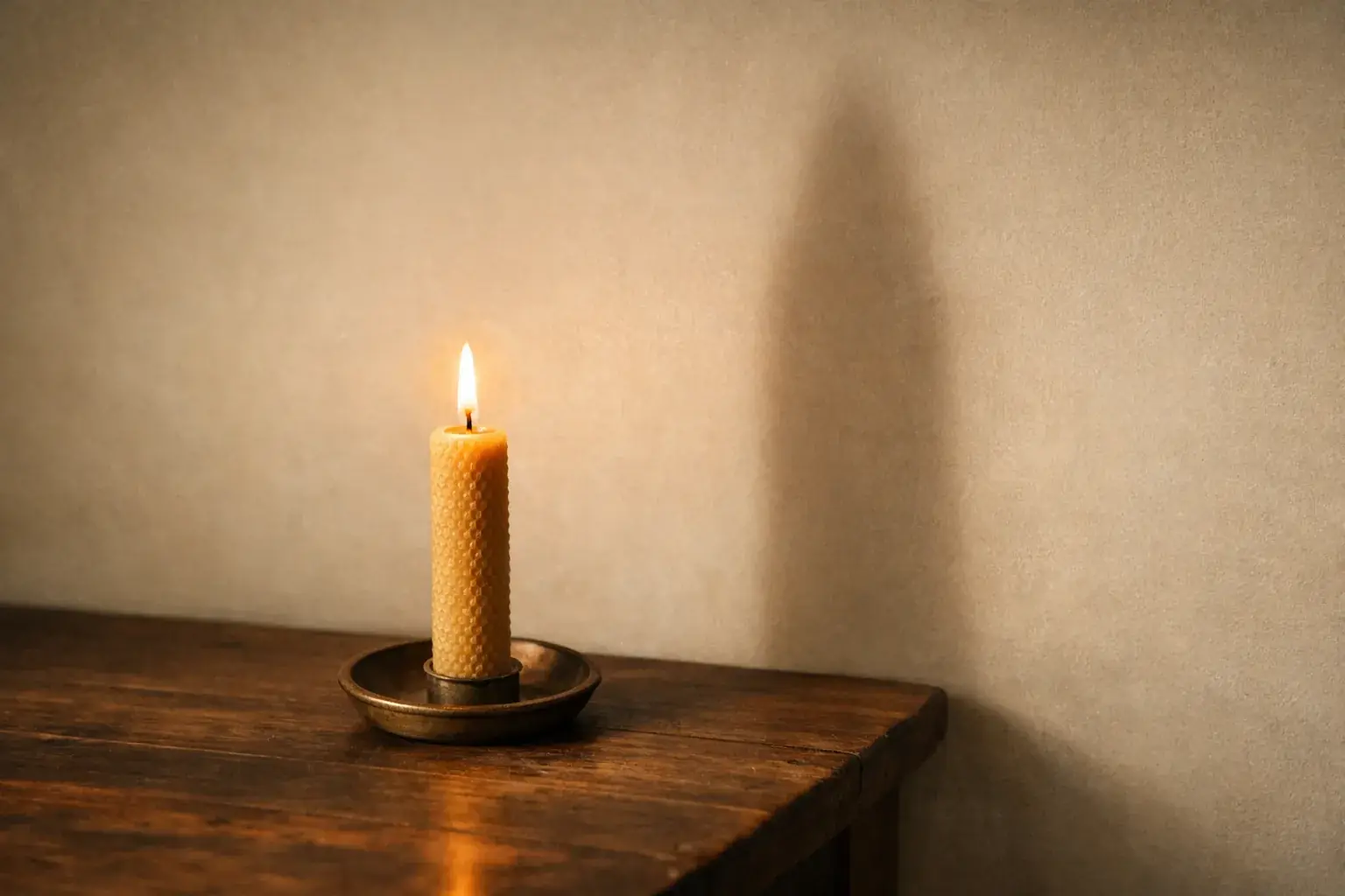 A honeycomb-textured beeswax candle burning in a brass dish on a dark wooden table, casting one soft-edged shadow on the pale wall beside it that is far taller than the candle itself.