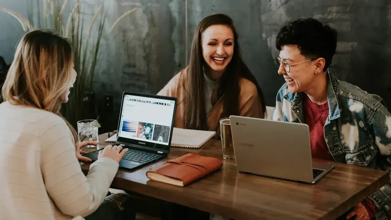 Three people laugh together while working on laptops at a wooden table in a casual indoor space.